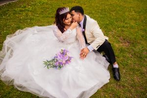 A couple in white wedding attire poses on green grass with purple flowers.