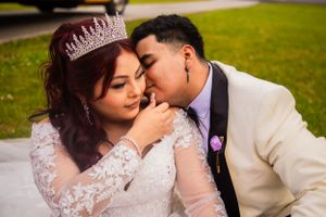 A couple in wedding attire shares an intimate moment on grass with purple flowers nearby.