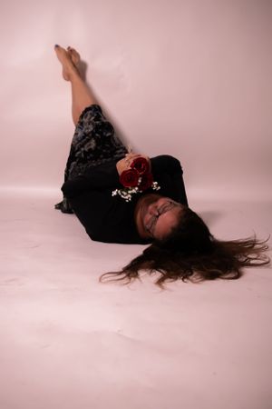 Person lying on pink studio floor holding red rose while wearing black outfit.