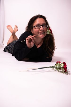 Person lying on white floor with red rose wearing black sweater.