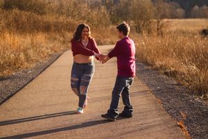 A couple in red tops and jeans hold hands while walking on a trail surrounded by autumn grass.