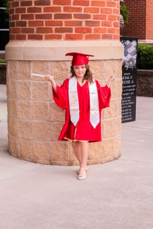 Graduate in bright red cap and gown poses against brick column outdoors.
