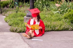 Graduate in red cap and gown sits on concrete path reading diploma.