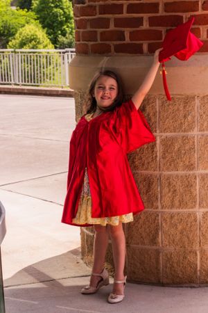 Excited graduate in red cap and gown raises arm in celebration by brick wall.
