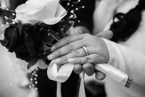 Close-up of wedding rings and hands holding white flowers with black accents.
