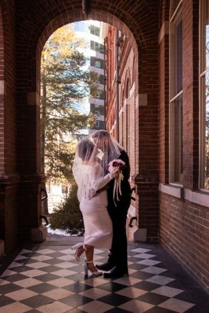 A couple embraces under a brick archway with checkered floors and autumn trees in background.