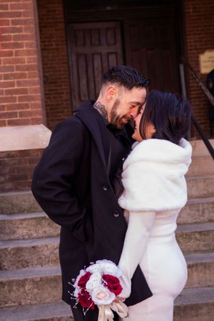 A winter wedding couple shares a tender moment on brick steps with a red and white bouquet.