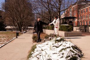 A winter wedding couple walking along a snowy path near brick buildings.
