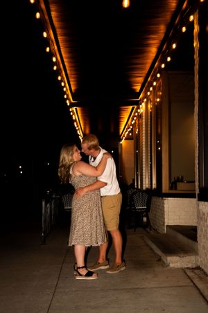 A couple embraces in a warmly lit outdoor corridor with string lights overhead.