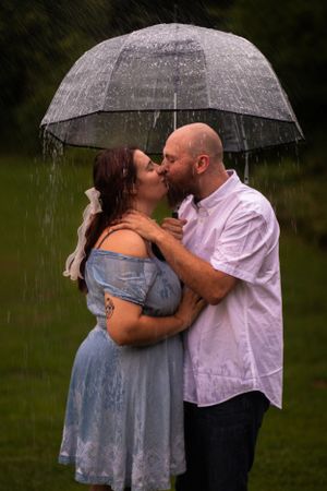 A couple kisses in the rain while sharing a clear umbrella in a romantic outdoor setting.