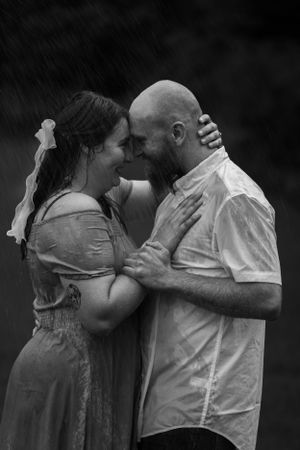 A couple embraces in the rain wearing light colored clothing in black and white photography.
