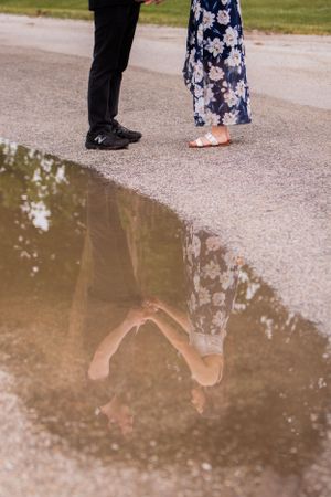 Reflection of a couple's feet in a puddle showing black shoes and a floral dress.