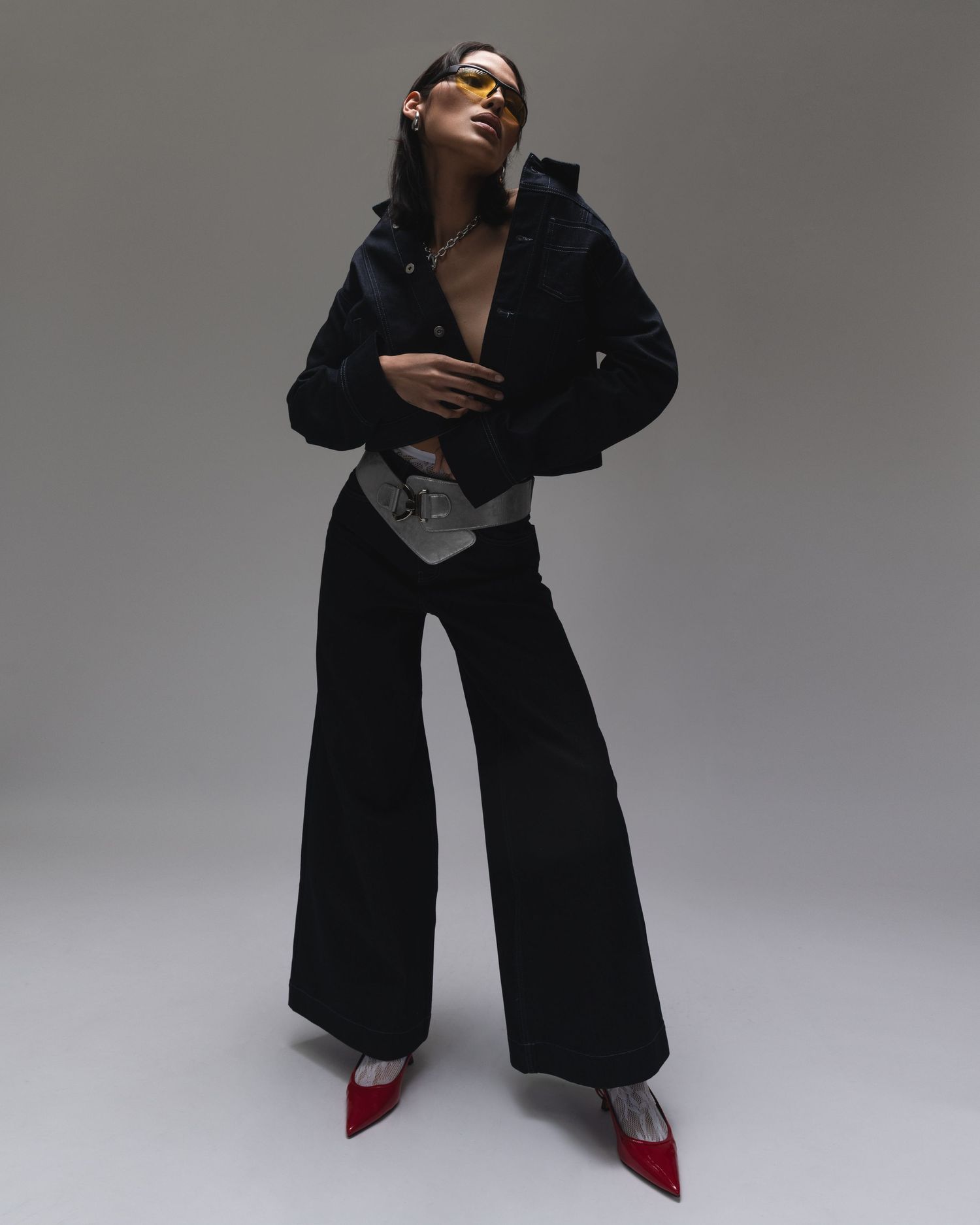 Fashion model in black wide-leg jumpsuit and red heels poses dramatically against gray studio backdrop.