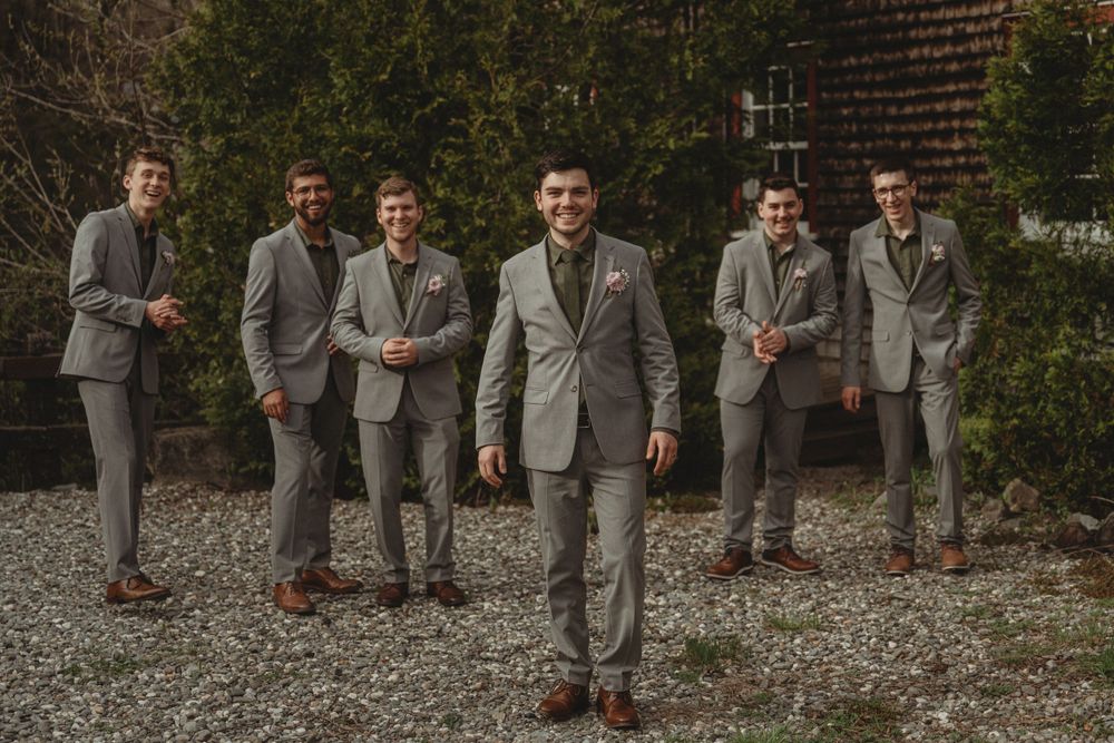Groomsmen in matching grey suits standing on gravel outside brick building.