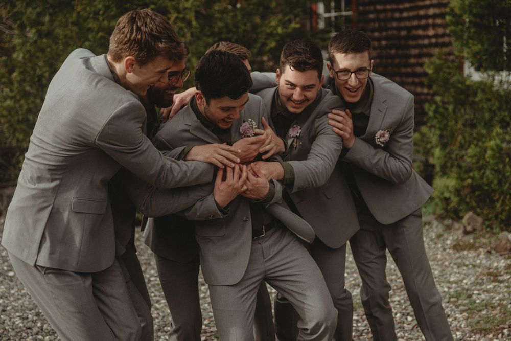 Playful groomsmen in grey suits huddled together in celebratory pose.