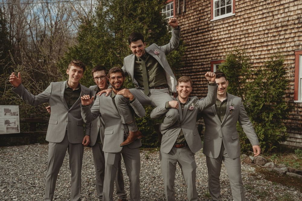 Groomsmen in matching gray suits pose playfully outside a brick building on a wedding day.