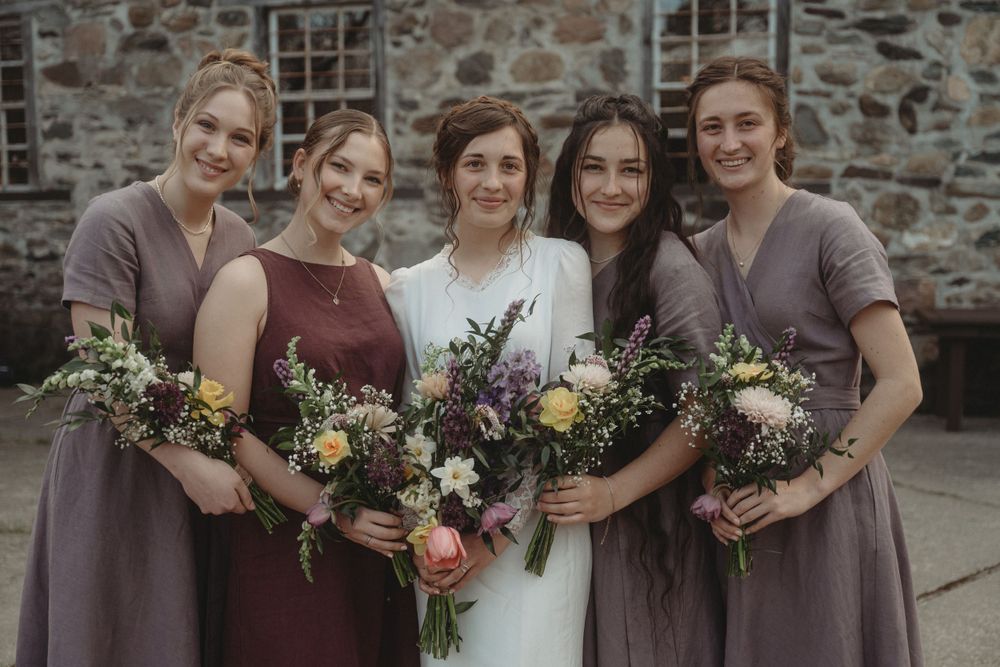 Bridesmaids in purple dresses hold colorful bouquets while posing with bride in white.