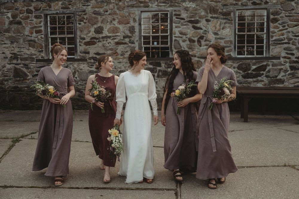 Wedding party in purple and white dresses walking along stone building exterior.
