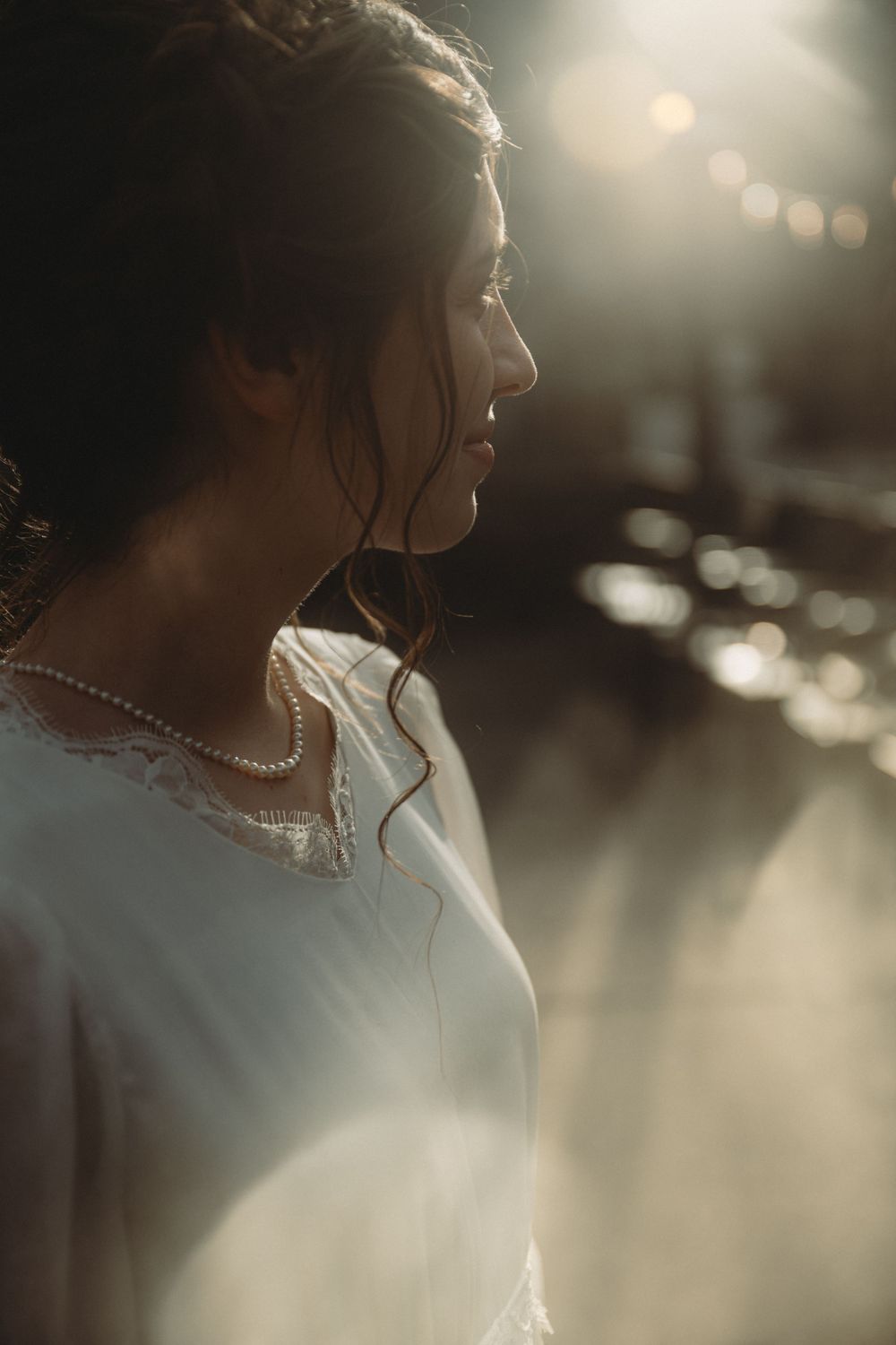 Side profile of bride in white dress with pearl necklace against evening light.