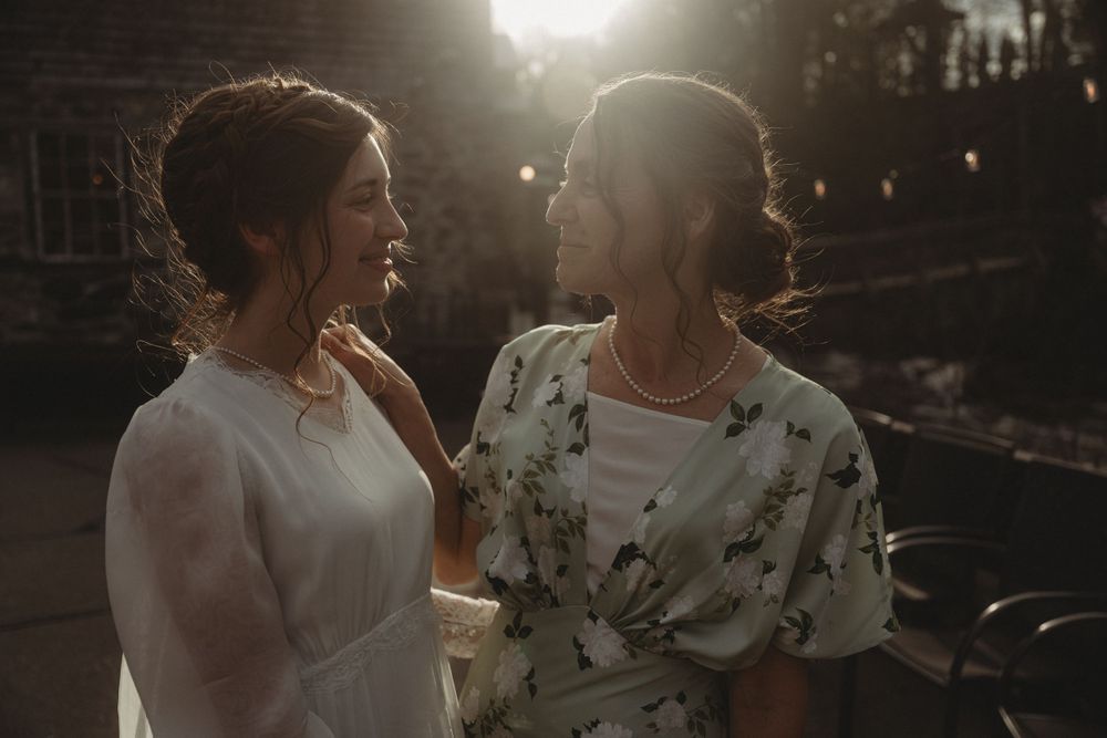 Two wedding guests share a joyful conversation at dusk with string lights glowing behind them.
