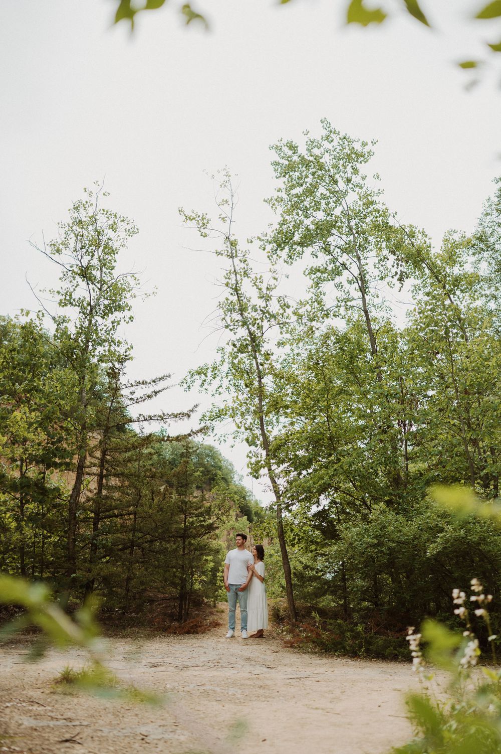 A figure stands on a serene dirt path surrounded by tall green trees and natural foliage on a cloudy day.