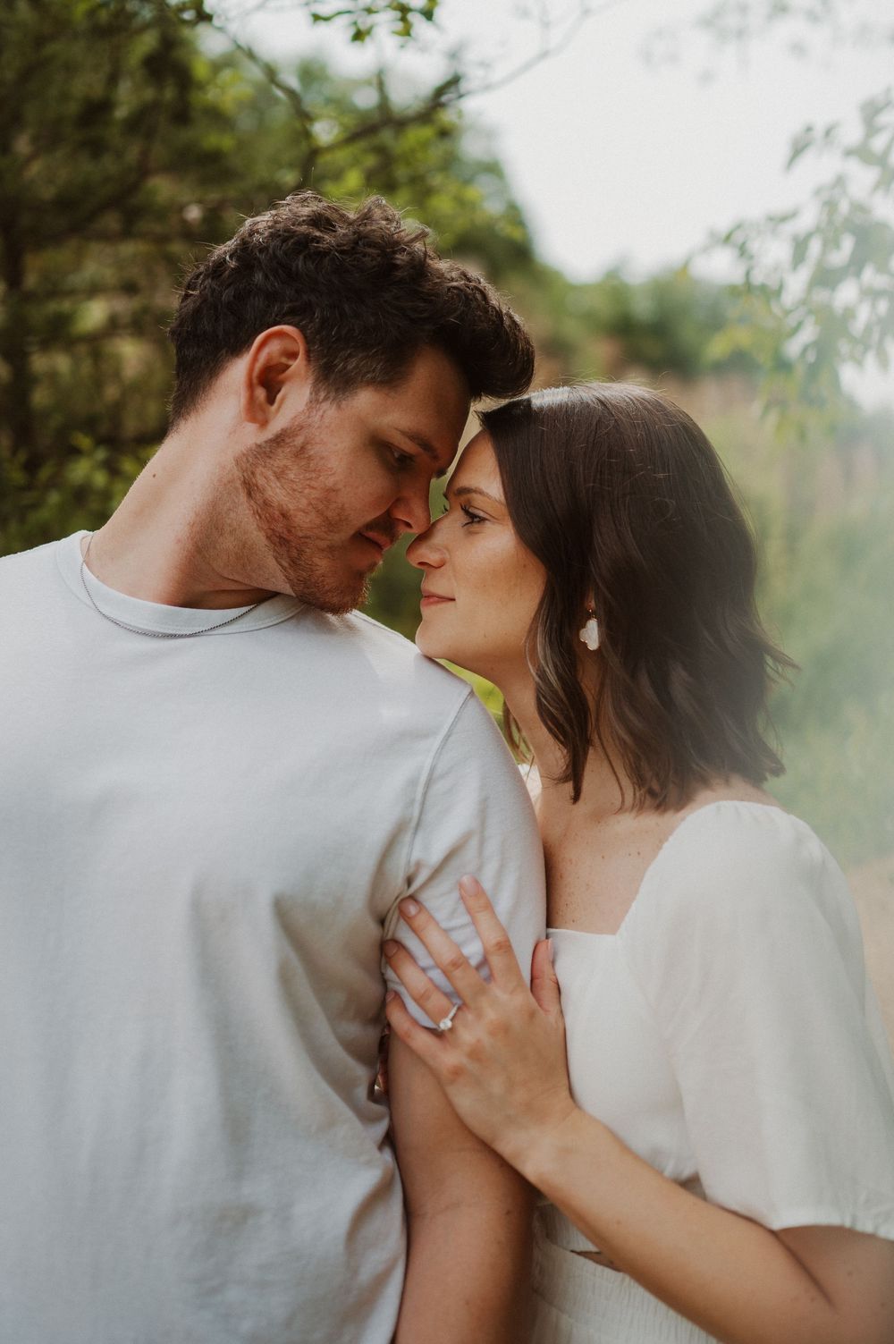 A romantic couple shares an intimate moment outdoors in soft natural lighting while wearing white casual clothing.