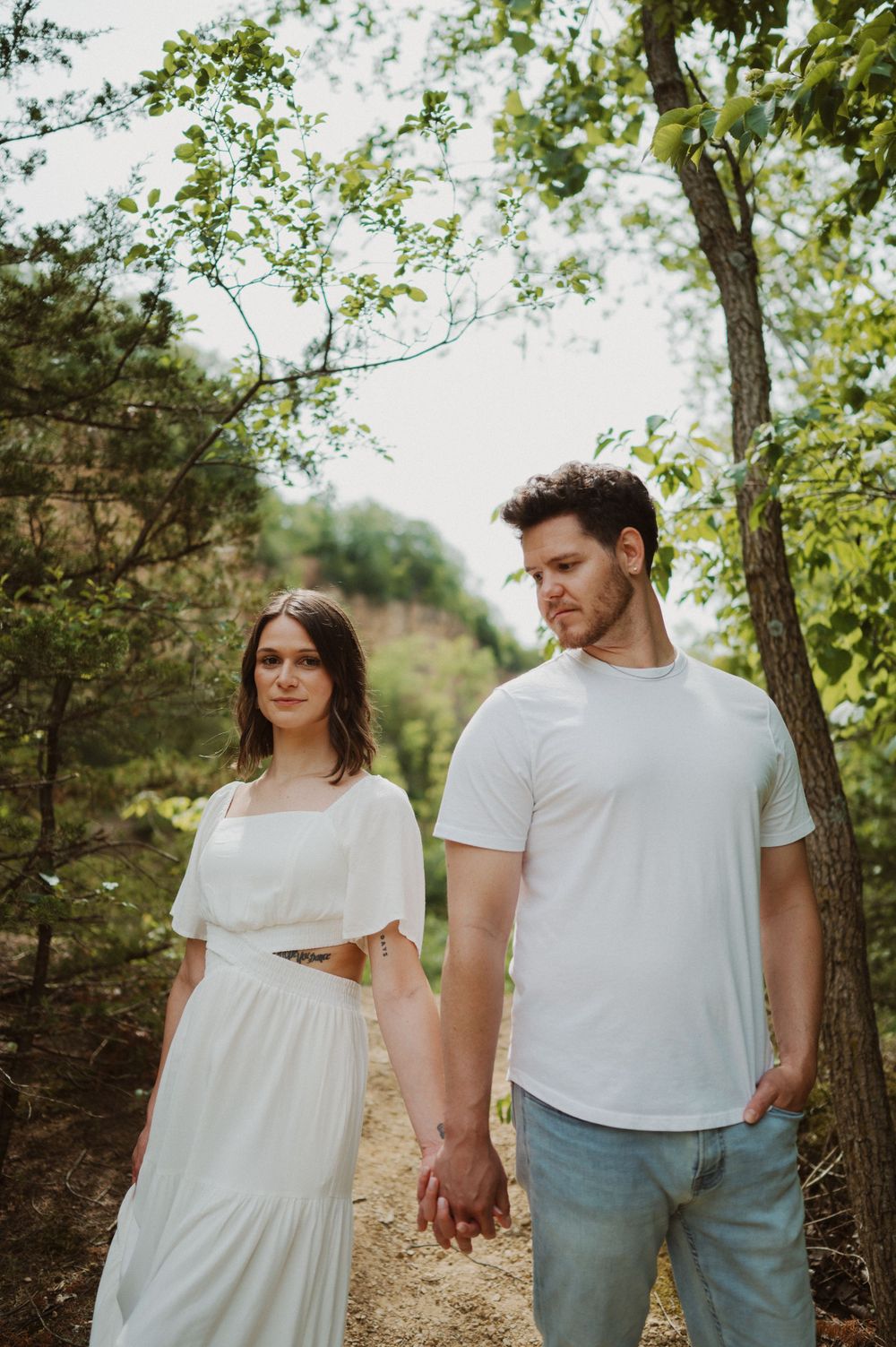 A couple in white outfits holding hands while walking on a wooded path in natural sunlight.