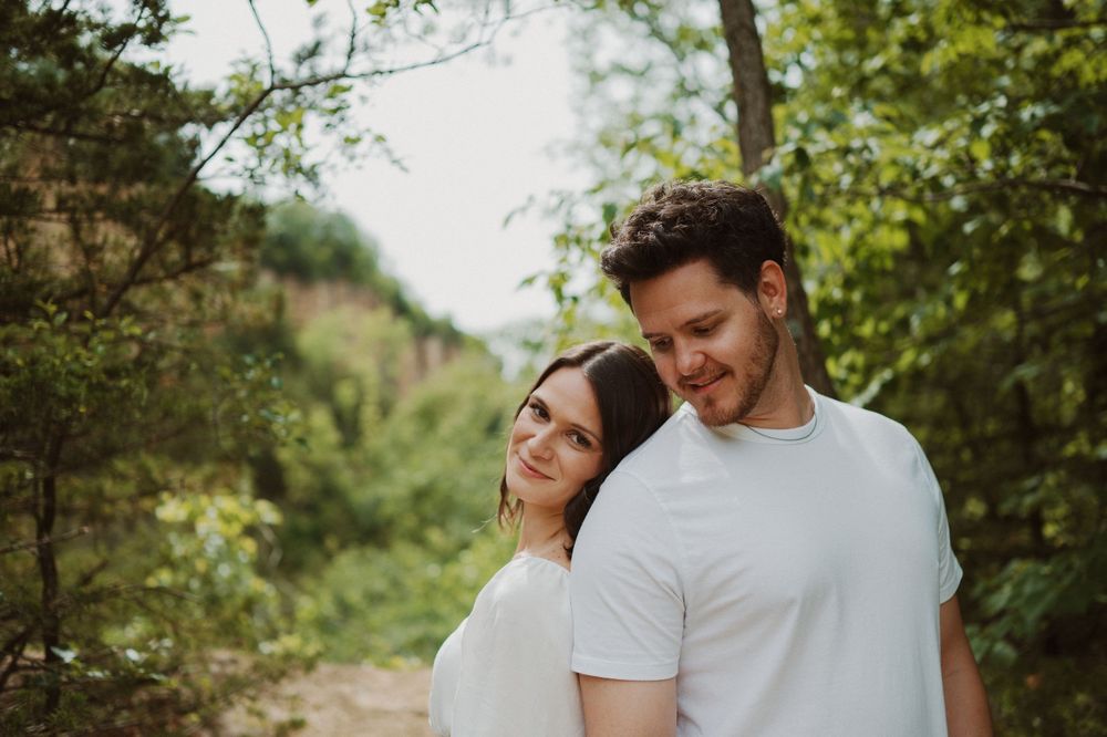 A couple shares an intimate moment while walking through a lush wooded area on a sunny day.