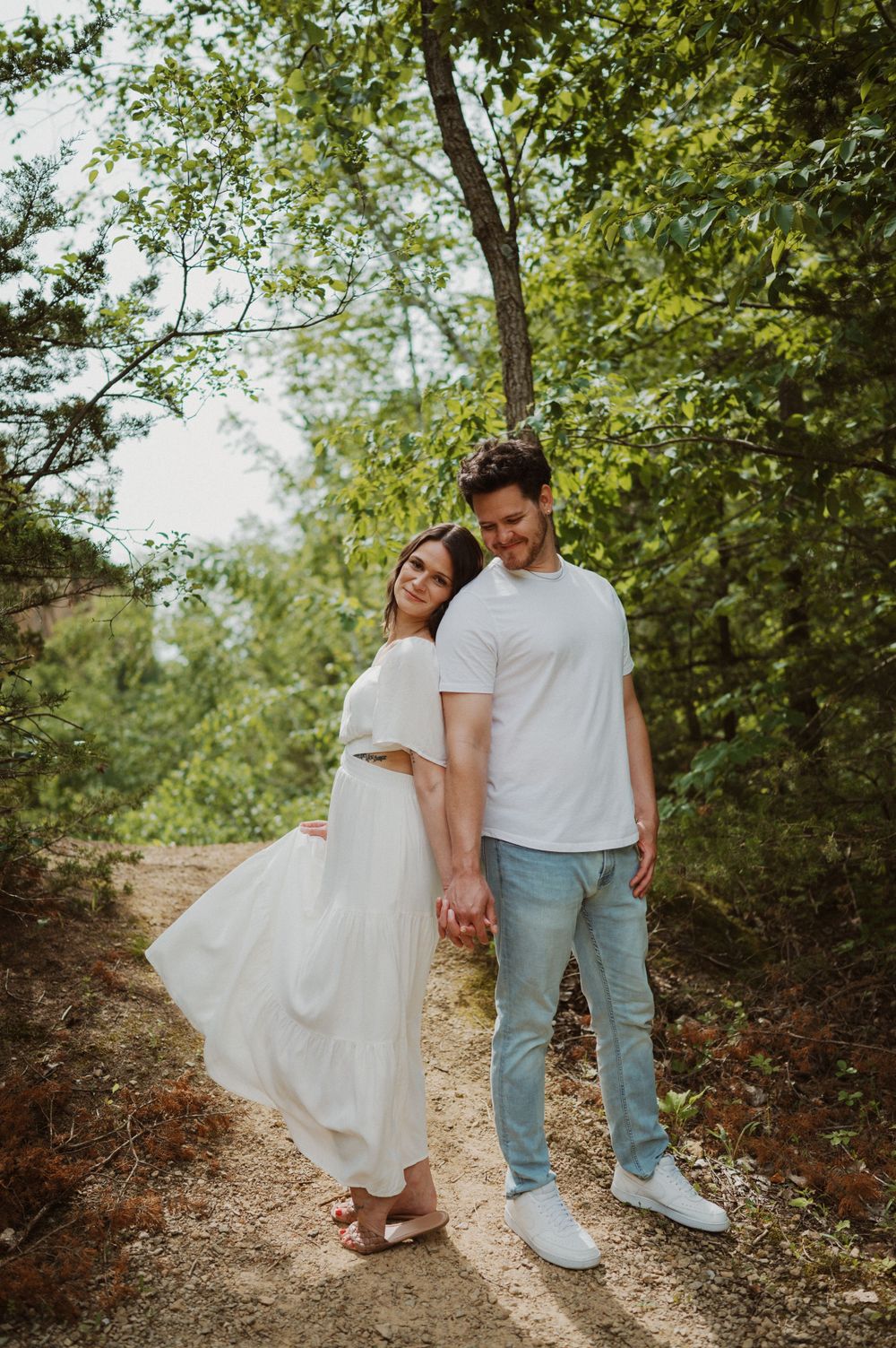 Romantic outdoor photo of a couple in white clothing standing together on a wooded path.