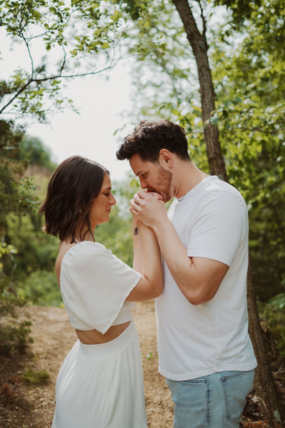Romantic couple in white outfits sharing an intimate moment on a sunny forest path.