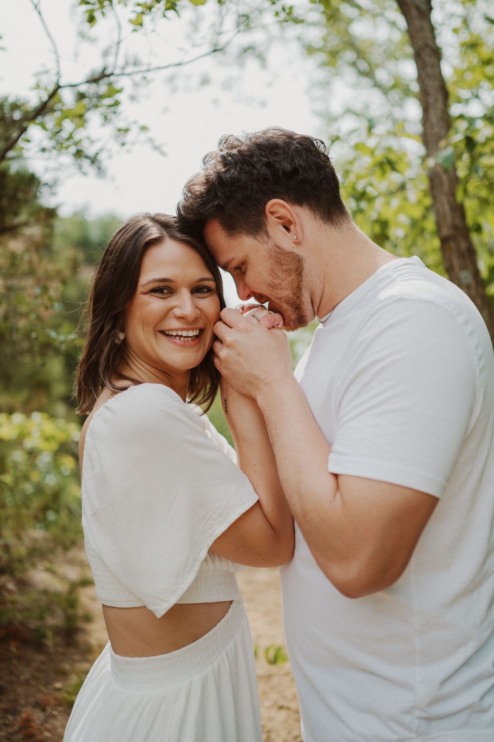Joyful couple in white clothing sharing laughs and embraces in a sunlit forest setting.