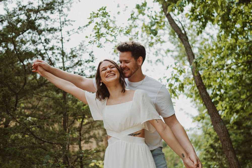 A joyful couple laughing and embracing in a sun-dappled forest setting with green foliage all around them.