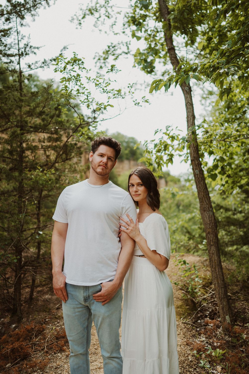 A romantic outdoor portrait of a couple in white clothing standing together in a wooded natural setting on a sunny day.