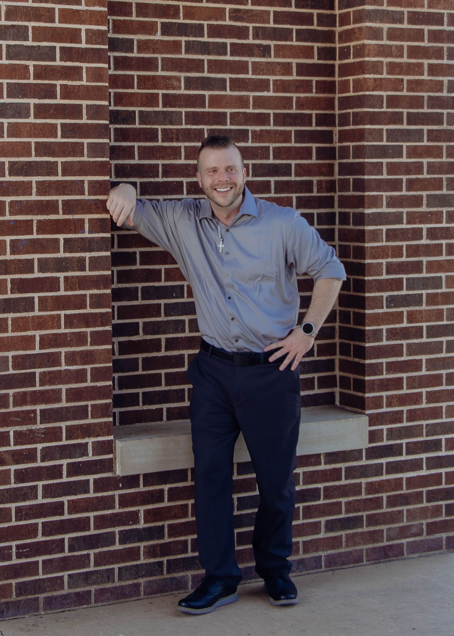 Someone posing casually against a brick wall while dressed in a gray button-down shirt and dark pants.