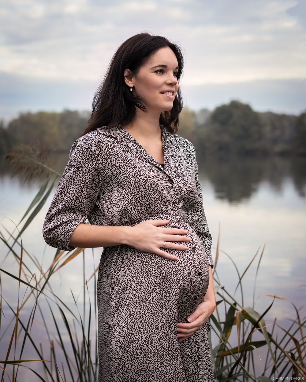 zwangere vrouw staat voor vijver met handen op haar buik tijdens zwangerschapsfotoshoot. gefotografeerd in beneden-leeuwen, maas&waal