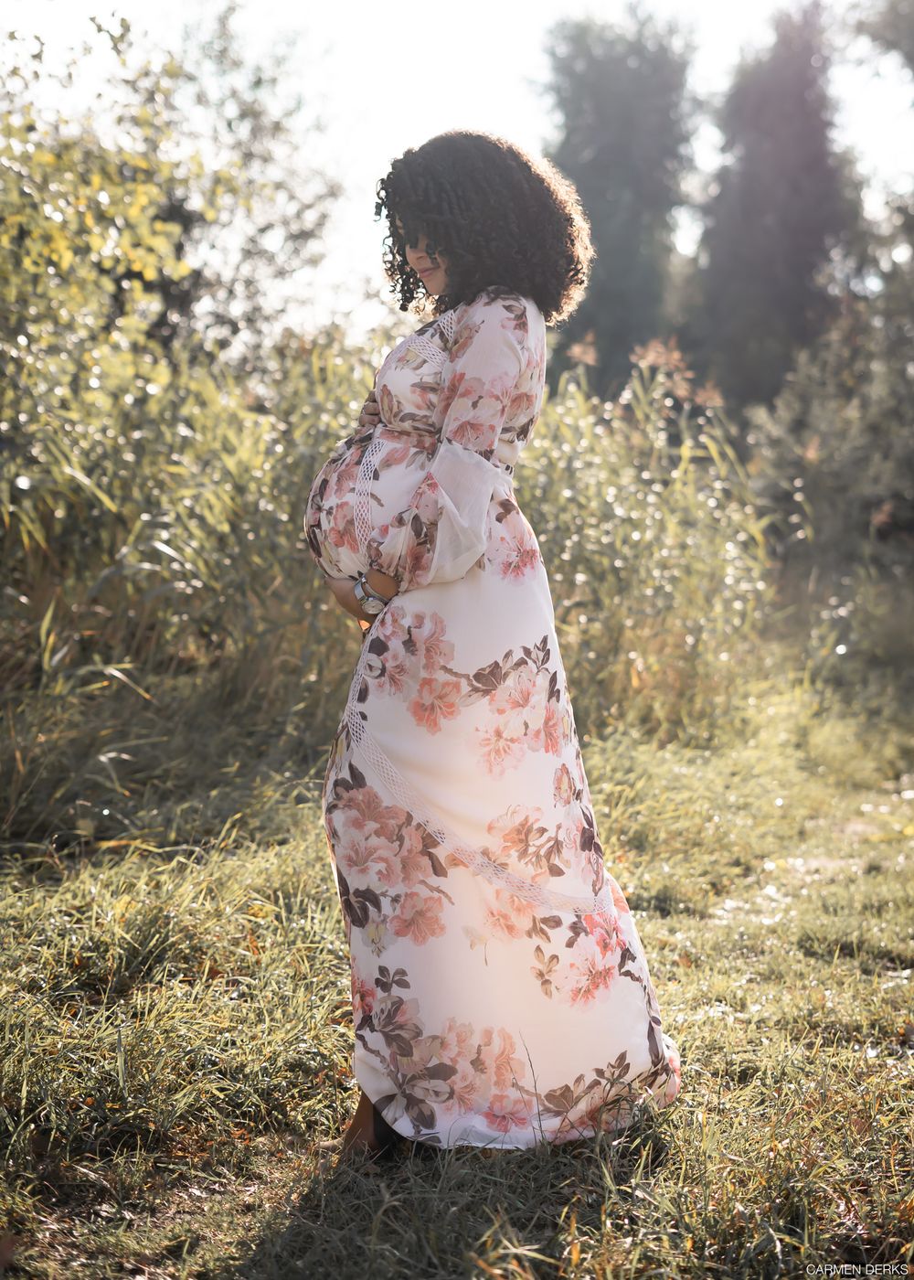 zwangere vrouw met luchtige bloemenjurk staat model in park met laag tegenlicht. gefotografeerd in Beneden-leeuwen, maas en waal.