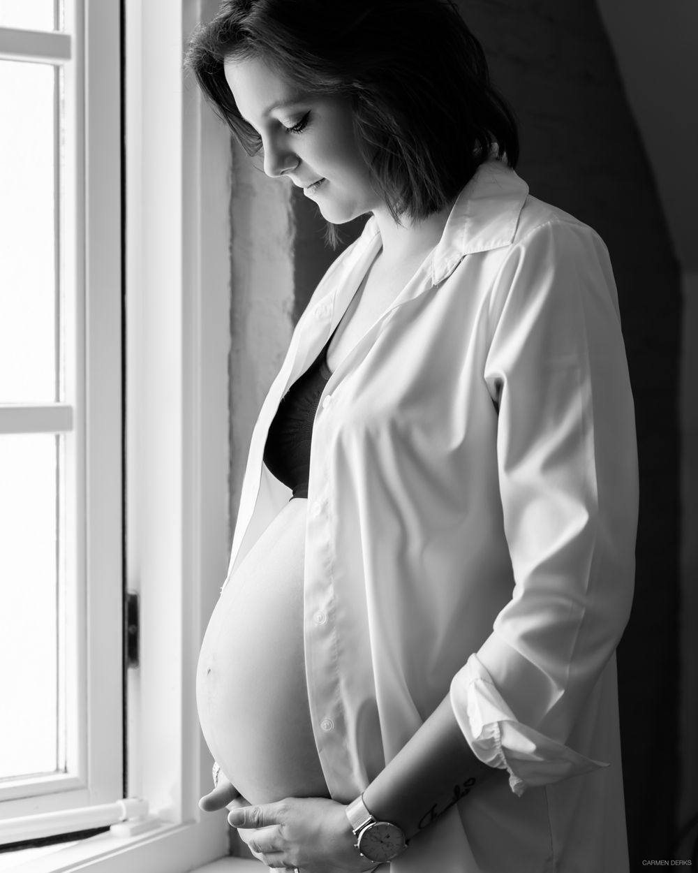 zwangere vrouw tijdens haar zwangerschapsshoot in huiselijke omgeving voor raam met blouse en ondergoed aan, zwart wit foto.