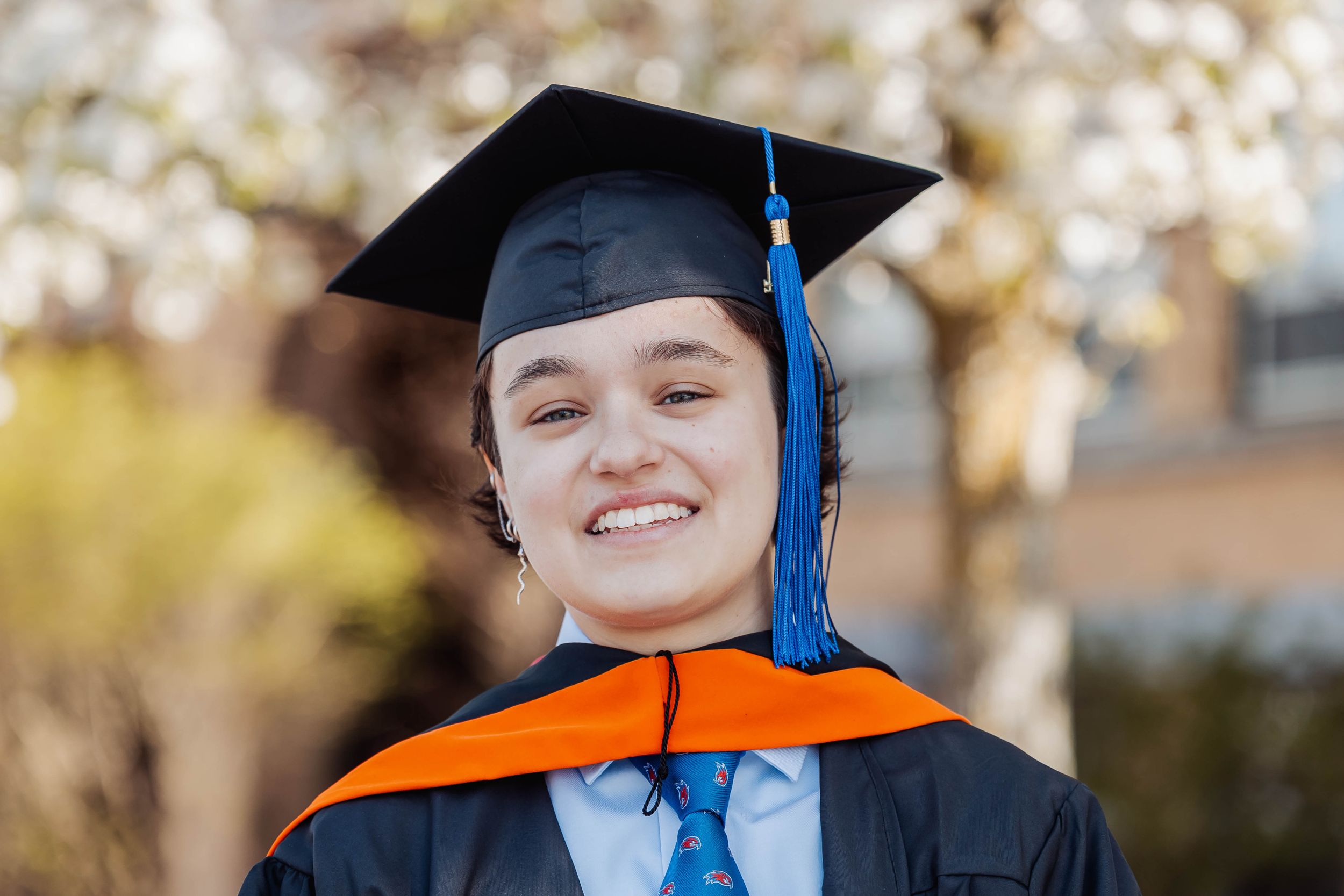 Graduate in black cap and gown with orange stole poses outdoors during spring.