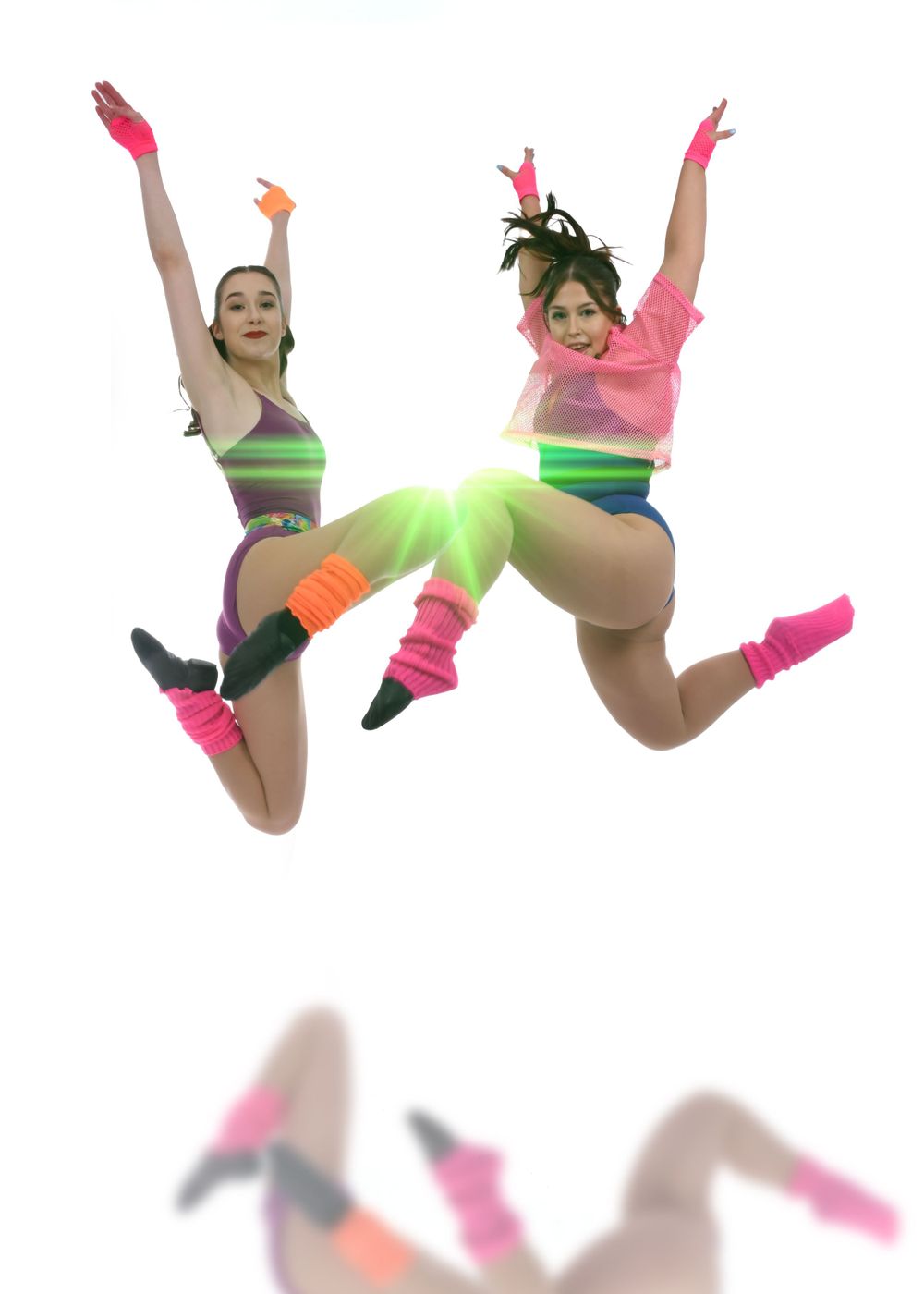 Two dancers in neon workout attire leap energetically with their arms raised and legs bent in a synchronized pose.