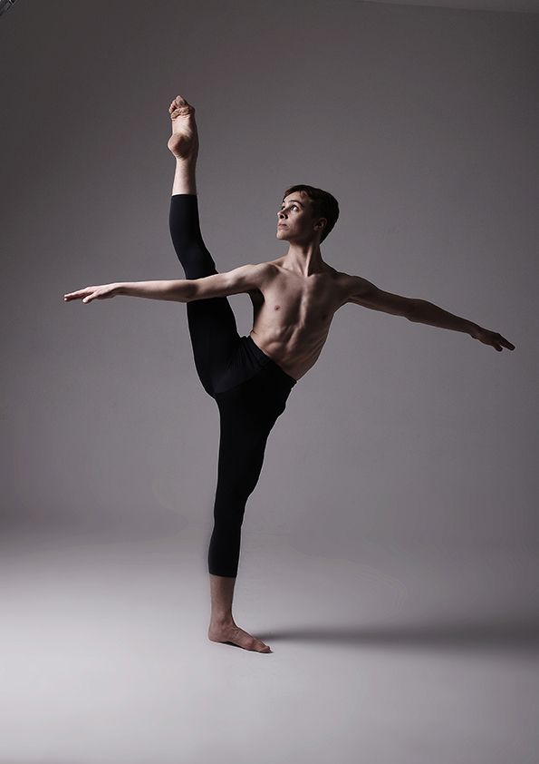 Contemporary dancer performs dramatic high leg extension pose in black pants against gray background.
