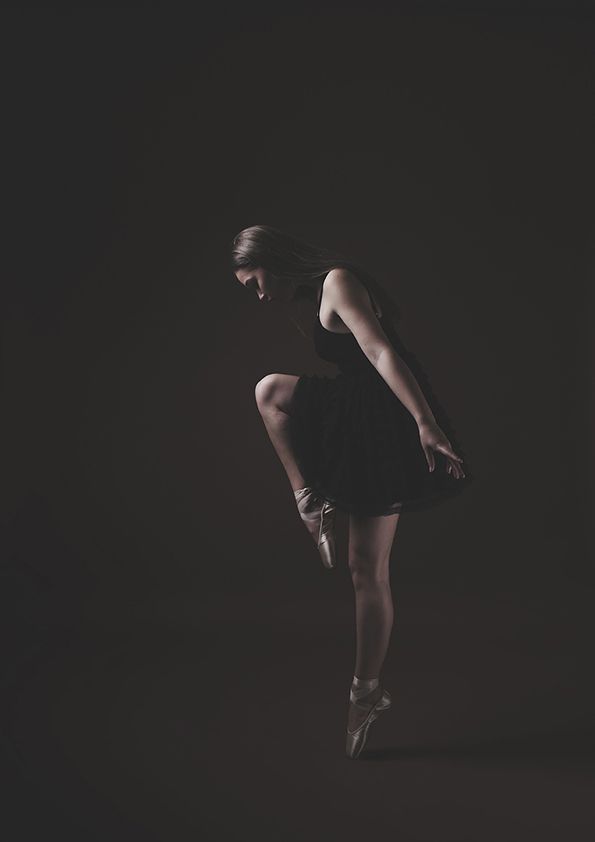 Moody low-key portrait of ballet dancer en pointe in black outfit against dark background.