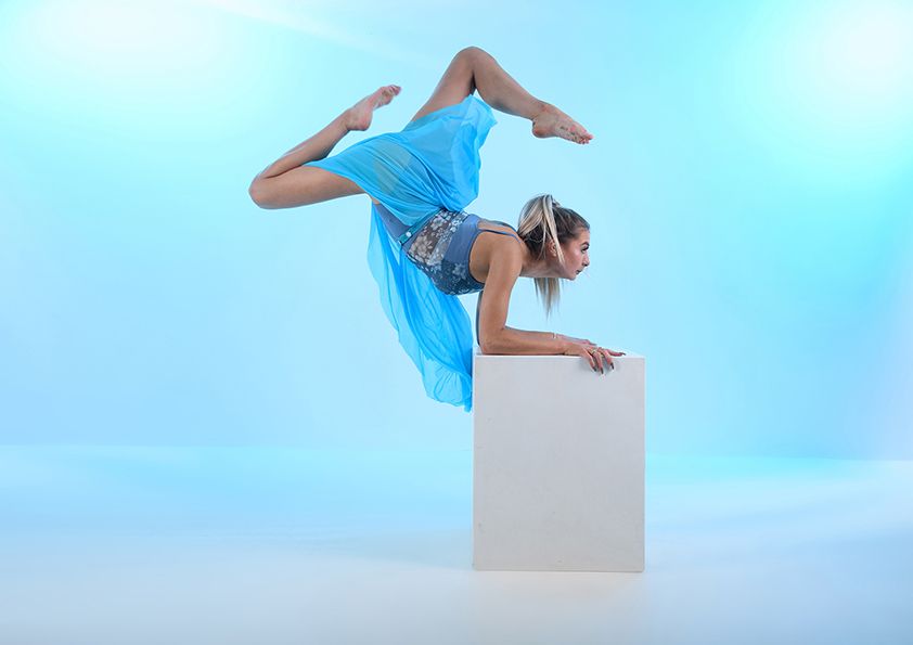 Athletic dancer in blue costume performs contortion pose on white pedestal against blue backdrop.