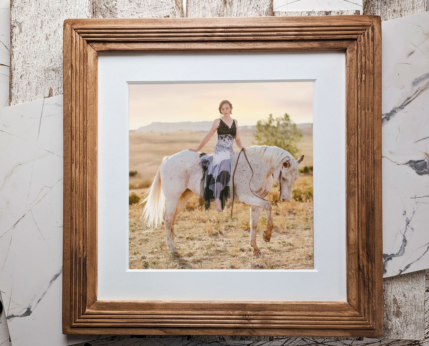 A framed rustic photograph of someone in a blue dress sitting on a white horse in a golden field during sunset.