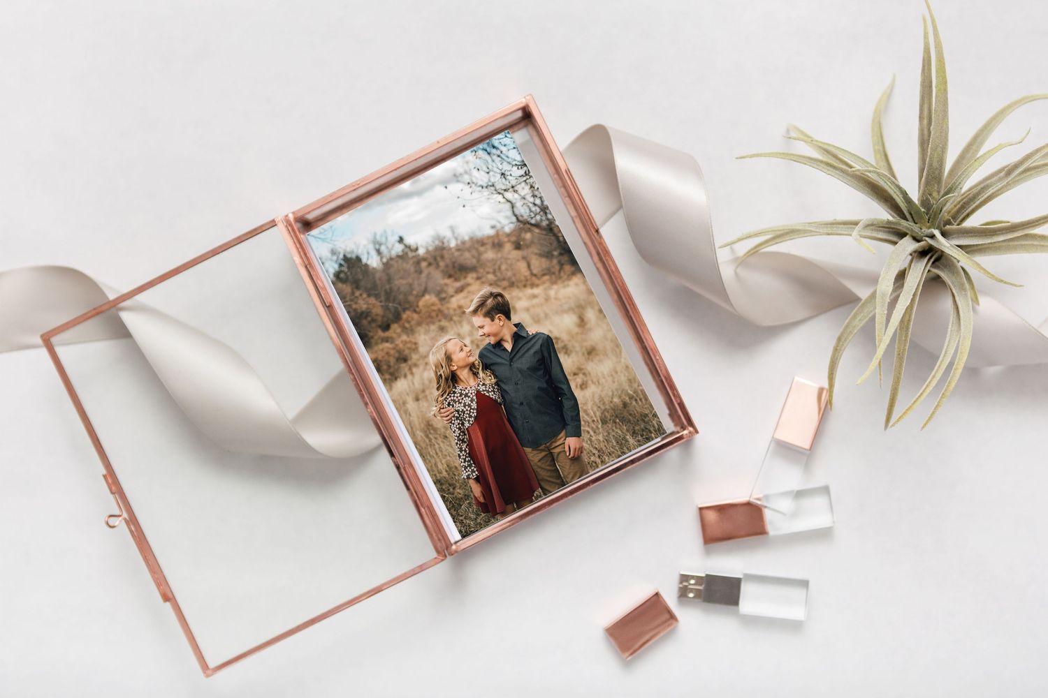 A rose gold photo album displaying an outdoor photograph lies open next to nail polish bottles and an air plant.
