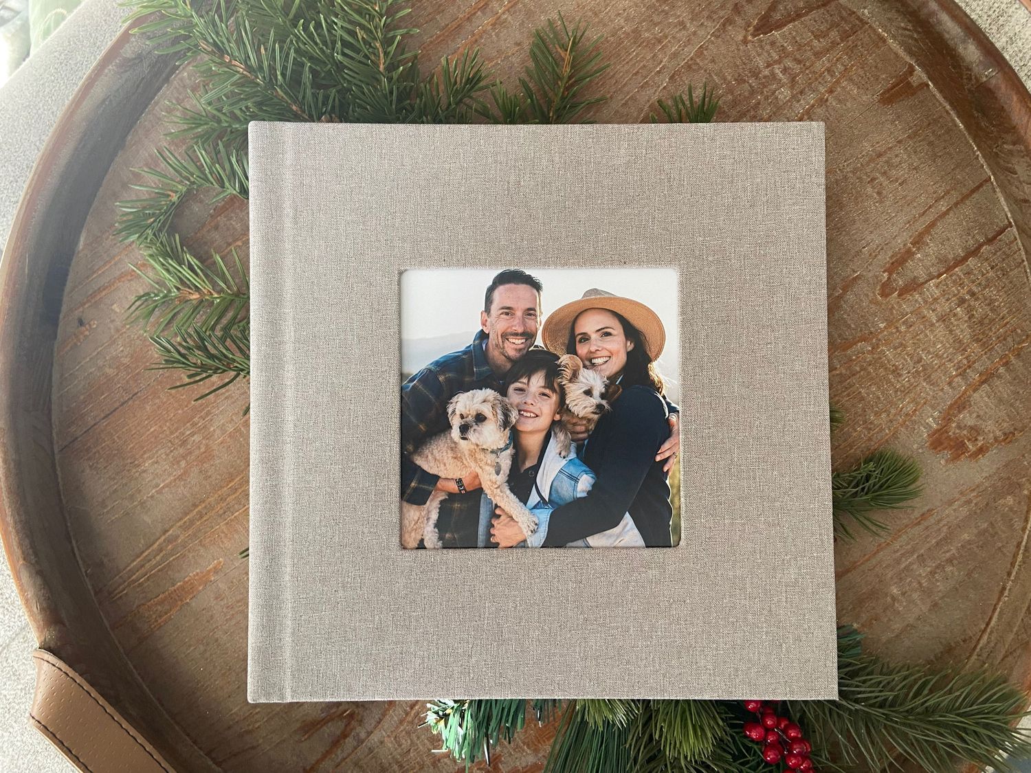 A beige linen photo album rests on a wooden tray decorated with evergreen branches and red berries.