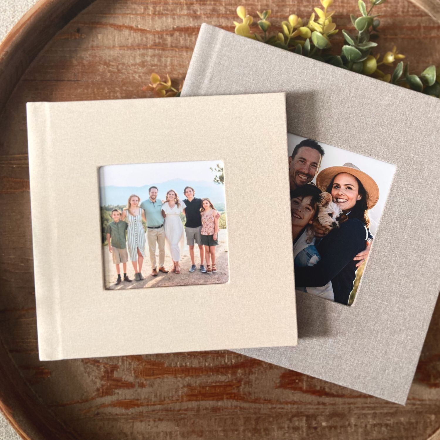 Two printed photo albums with memories displayed on a round wooden table with decorative greenery.