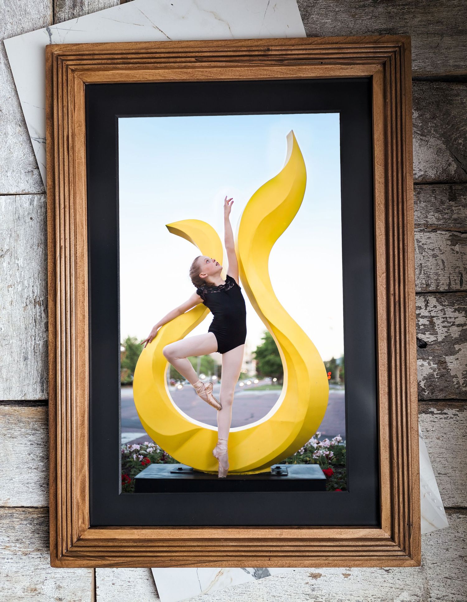 A wooden framed photo shows a dancer performing on a curved yellow artistic sculpture outdoors.
