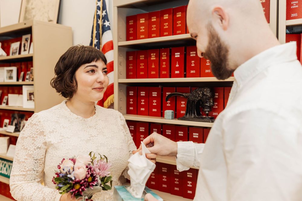 A couple shares a tender moment during ceremony with red bookshelves and flag in background.