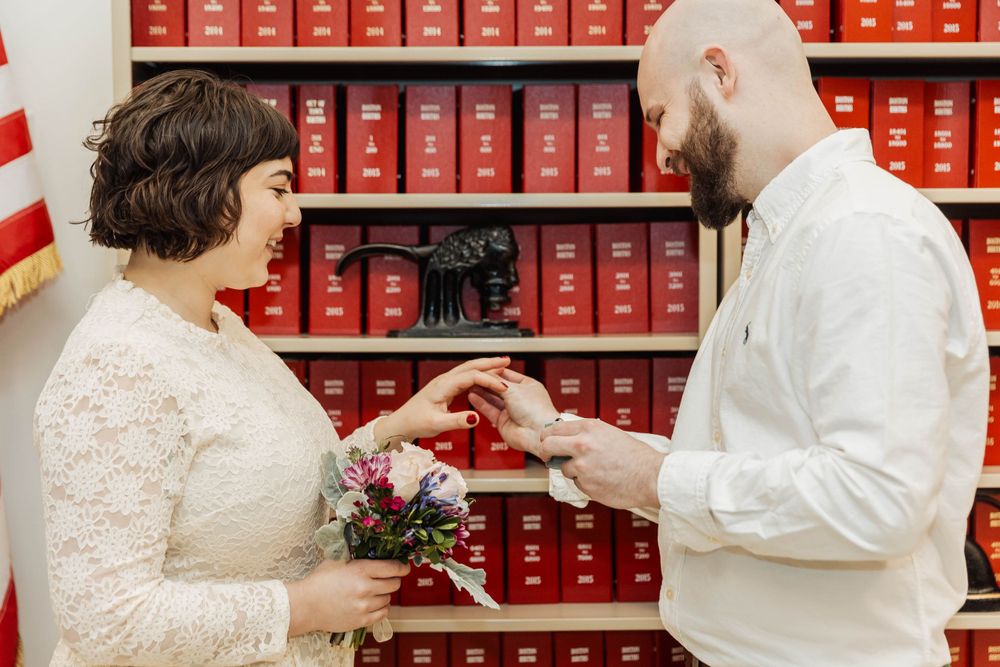 A couple exchanges rings during ceremony in front of red bookshelves with American flag.