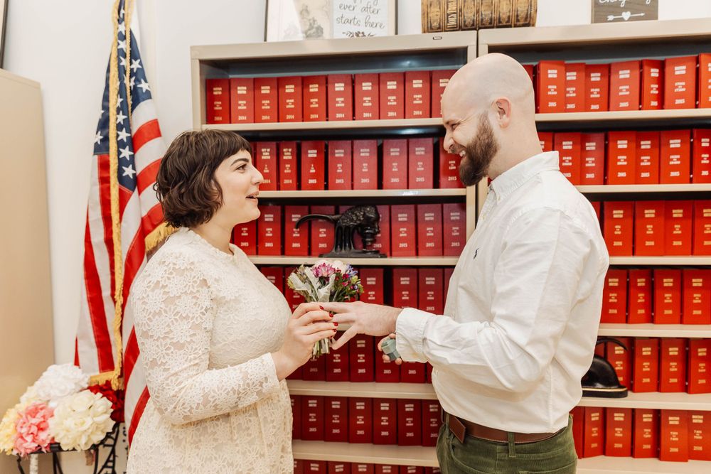 A couple shares vows during ceremony in front of red law books and American flag.
